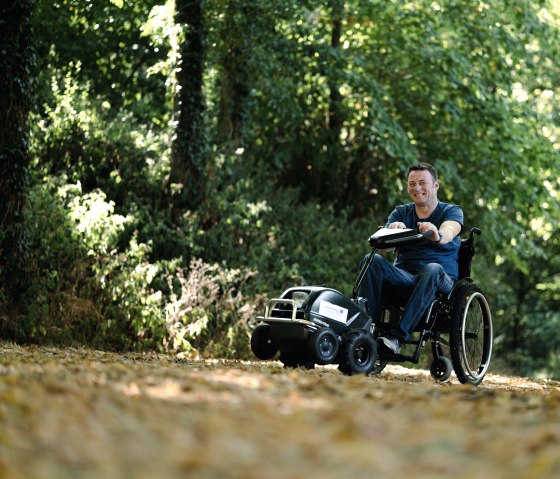 Personne en fauteuil roulant avec un appareil de traction pour fauteuil roulant sur un sentier de randonnée dans le parc naturel Südeifel, © Naturpark Südeifel, Thomas Urbany